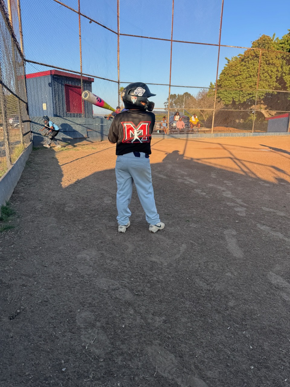 A 9U player in a black jersey with the red M and a batting helmet, bat over his shoulder, waiting on deck